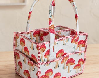 May include: A rectangular fabric storage caddy featuring a white background with a repeating red and orange mushroom pattern. The organizer has two upright handles, quilted stitching, and red and white striped fabric trim along the edges.