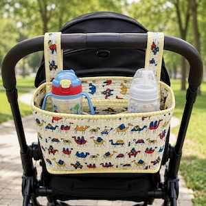 May include: A fabric pram organiser featuring a cream background with a repeating pattern of colourful camels in red, blue, and yellow. The organiser has two vertical straps for attachment and contains a plastic water bottle and a baby bottle.