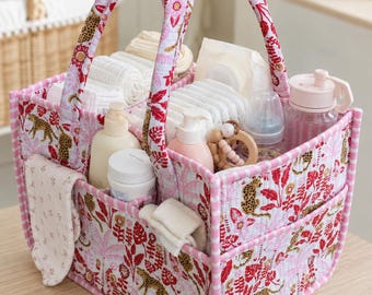 May include: A rectangular quilted fabric storage caddy featuring a pink and red pattern of leopards and tropical foliage. The organizer has two sturdy handles, multiple exterior pockets, and internal dividers filled with baby care items.