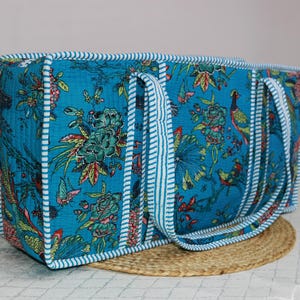 May include: A rectangular quilted tote bag in a vibrant turquoise blue fabric featuring a multi-colored floral and bird print. The bag is finished with white and blue striped fabric piping along all edges and includes two matching striped handles.