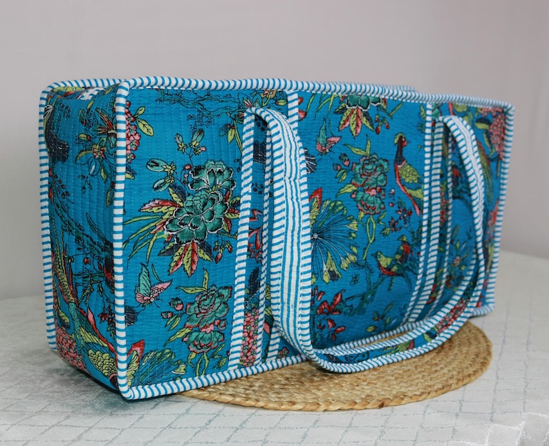 May include: A rectangular quilted tote bag in a vibrant turquoise blue fabric featuring a multi-colored floral and bird print. The bag is finished with white and blue striped fabric piping along all edges and includes two matching striped handles.
