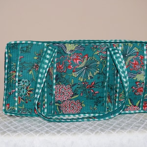 May include: A rectangular quilted tote bag in a deep teal color featuring a vibrant floral pattern in shades of red, pink, and green. The bag is finished with a white and teal checkered trim along the edges and the two shoulder straps.