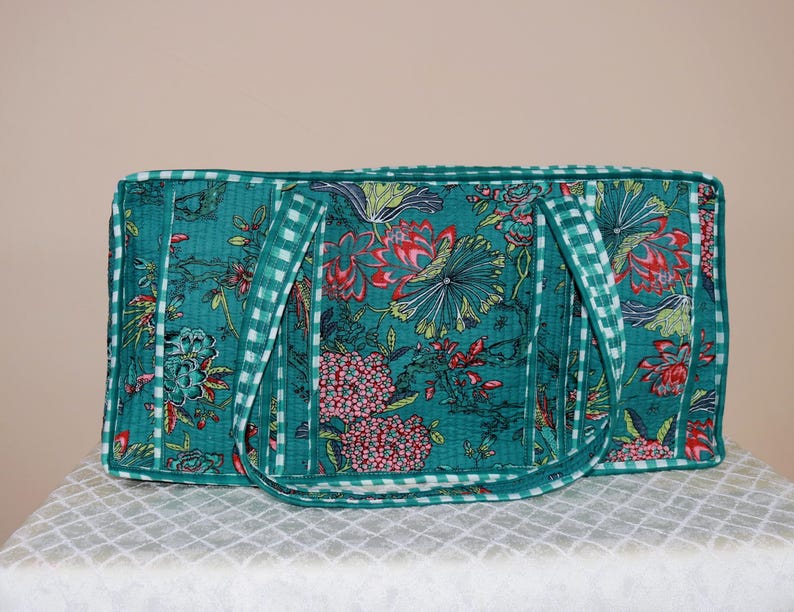 May include: A rectangular quilted tote bag in a deep teal color featuring a vibrant floral pattern in shades of red, pink, and green. The bag is finished with a white and teal checkered trim along the edges and the two shoulder straps.