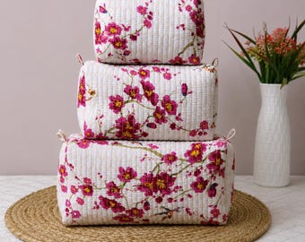 May include: A set of three rectangular quilted fabric pouches stacked vertically. Each pouch is off-white with a repeating pattern of vibrant magenta and pink cherry blossom flowers on thin brown branches, featuring vertical stitching throughout.