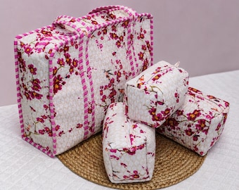 May include: A quilted fabric set consisting of one large rectangular tote bag and three smaller matching zippered pouches. Each item features a cream-colored base with a repeating pattern of vibrant pink cherry blossoms and thin pink striped trim.