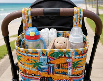 May include: A quilted fabric stroller organizer featuring a vibrant tropical beach print with palm trees, surfboards, and a cafe. The bag hangs from the stroller handle and contains a plastic water bottle, a baby bottle, and a plush rabbit toy.