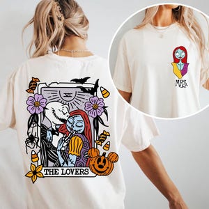 May include: An off-white t-shirt featuring a back graphic of a tarot card titled THE LOVERS with Jack and Sally characters, surrounded by pumpkins, bats, and flowers. The front chest area displays a small graphic of Sally and the text MRS.