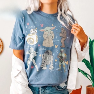K&ouml;nnte beinhalten: Ein blaues Kurzarm-T-Shirt mit einem Grafikdruck von Star Wars-Charakteren, darunter BB-8, Baby Yoda, Chewbacca, ein Stormtrooper, R2-D2 und Boba Fett, akzentuiert durch kleine rosa und gelbe Mickey-Maus-Kopf-Silhouetten und wei&szlig;e Sterne.