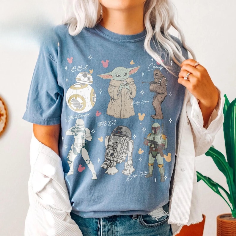 K&ouml;nnte beinhalten: Ein blaues Kurzarm-T-Shirt mit einem Grafikdruck von Star Wars-Charakteren, darunter BB-8, Baby Yoda, Chewbacca, ein Stormtrooper, R2-D2 und Boba Fett, akzentuiert durch kleine rosa und gelbe Mickey-Maus-Kopf-Silhouetten und wei&szlig;e Sterne.