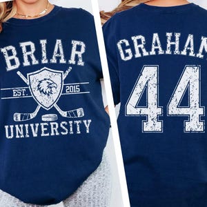 May include: A navy blue t-shirt featuring a distressed white graphic on the front that reads BRIAR UNIVERSITY EST. 2015 with an eagle shield and hockey sticks. The back of the shirt displays the name GRAHAM and the number 44 in distressed white text.