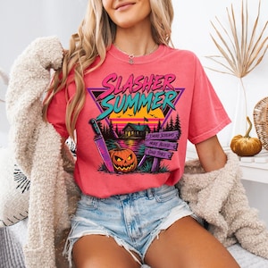 May include: A coral-coloured t-shirt featuring a graphic print with the text Slasher Summer, a cabin, a jack-o'-lantern, a knife, and a sign that reads S'more Screams, More Blood, Good Times, set against a sunset and a dark forest background.