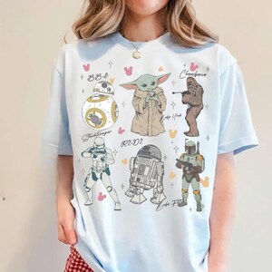 K&ouml;nnte beinhalten: Ein hellblaues T-Shirt mit einem Grafikdruck von sechs Star Wars-Charakteren, darunter BB-8, Baby Yoda, Chewbacca, ein Stormtrooper, R2-D2 und Boba Fett. Das Design enth&auml;lt kleine rosa und orangefarbene mausf&ouml;rmige Symbole sowie silberne Sternakzente.