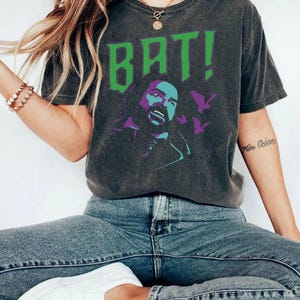 May include: A charcoal gray short-sleeve t-shirt featuring a graphic print of a person with fangs and the text BAT! in large green letters. The design includes purple silhouettes of flying bats and a stylized portrait of a person with a beard.