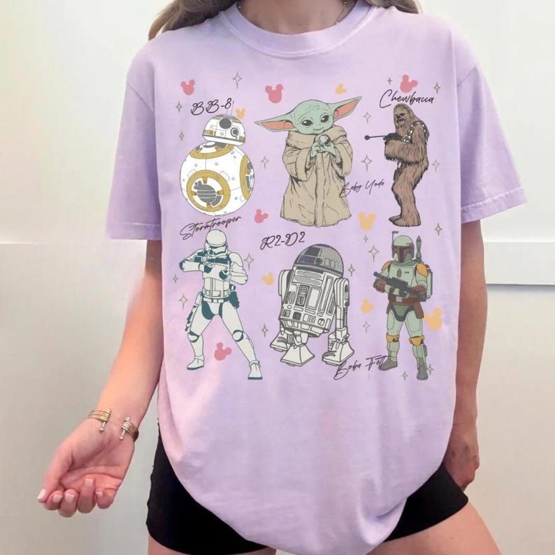 K&ouml;nnte beinhalten: Ein hellviolettes T-Shirt mit einem Grafikdruck von sechs Star Wars-Charakteren: BB-8, Baby Yoda, Chewbacca, ein Stormtrooper, R2-D2 und Boba Fett. Das Design enth&auml;lt kleine Stern- und Mausohr-Symbole, die zwischen den Illustrationen verteilt sind.