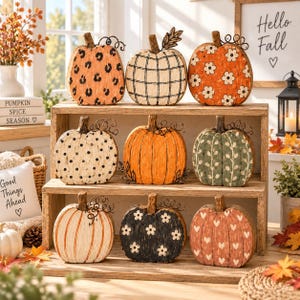 May include: A collection of nine decorative wooden pumpkins arranged on a three-tiered wooden shelf. Each pumpkin features a unique pattern, including leopard print, plaid, floral, polka dot, solid orange, vine, striped, and heart designs.