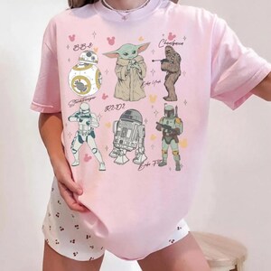 K&ouml;nnte beinhalten: Ein hellrosa T-Shirt mit einem Grafikdruck von Star Wars-Charakteren, darunter BB-8, Baby Yoda, Chewbacca, ein Stormtrooper, R2-D2 und Boba Fett, begleitet von kleinen gelben und rosa mausf&ouml;rmigen Symbolen und Sternakzenten auf der Vorderseite.