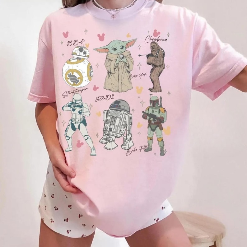 K&ouml;nnte beinhalten: Ein hellrosa T-Shirt mit einem Grafikdruck von Star Wars-Charakteren, darunter BB-8, Baby Yoda, Chewbacca, ein Stormtrooper, R2-D2 und Boba Fett, begleitet von kleinen gelben und rosa mausf&ouml;rmigen Symbolen und Sternakzenten auf der Vorderseite.