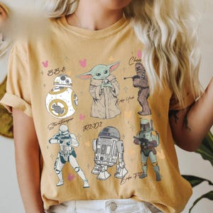 K&ouml;nnte beinhalten: Ein senfgelbes T-Shirt mit einem Grafikdruck von Star Wars-Figuren, darunter BB-8, Baby Yoda, Chewbacca, ein Stormtrooper, R2-D2 und Boba Fett, erg&auml;nzt durch kleine rosa und orangefarbene Mauseohr-Silhouetten und Sternakzente.