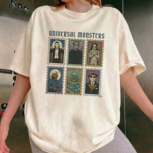 May include: A cream-colored short-sleeve t-shirt featuring a graphic print of six postage stamp-style illustrations of Universal Monsters characters, with the text "UNIVERSAL MONSTERS" printed in a dark font above the grid of character images.
