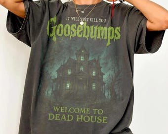May include: A charcoal gray short-sleeve t-shirt featuring a graphic print of a dark, spooky mansion. The text reads IT WILL JUST KILL YOU, Goosebumps, and WELCOME TO DEAD HOUSE in a distressed, light green font across the front of the shirt.
