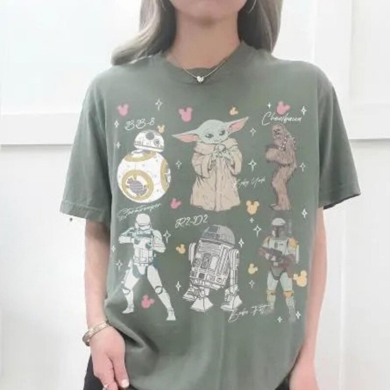 K&ouml;nnte beinhalten: Ein olivgr&uuml;nes Kurzarm-T-Shirt mit einem Grafikdruck von Star Wars-Charakteren, darunter BB-8, Grogu, Chewbacca, ein Stormtrooper, R2-D2 und Boba Fett, sowie kleinen rosa und gelben Mausohr-Symbolen und verstreuten Sternformen.