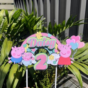 Pode incluir: Um topo de bolo de papel multicamadas com um arco-íris colorido, o nome Daisy em letras cor-de-rosa, o número 2 em cor-de-rosa e personagens da Peppa Pig. O design inclui pequenas flores amarelas, corações azuis e cor-de-rosa e balões.