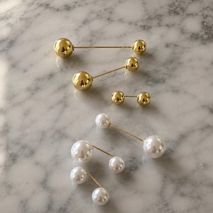 May include: A collection of six double-ended pins featuring polished gold-tone metal spheres and smooth white faux pearl spheres. Each pin consists of a thin metal bar connecting two spheres of varying sizes, arranged in a scattered formation.