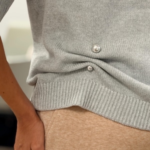 May include: A light gray knit sweater featuring short sleeves and a ribbed hem. The side of the garment is accented with two small, round, polished silver-tone metal buttons positioned vertically along the hip area.