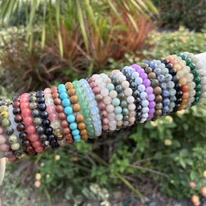 May include: A collection of beaded bracelets in various colors, including pink, black, blue, green, and purple. The bracelets are displayed on a white cylindrical stand against a backdrop of greenery. Each bracelet appears to be made of small, round beads.