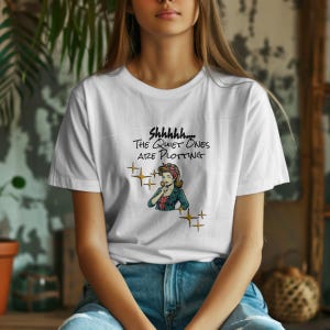 Puede incluir: Camiseta blanca con el texto "Shhhhh... The Quiet Ones Are Plotting" y un gráfico de una mujer con una blusa de lunares rojos y blancos y una chaqueta verde. También hay gráficos de estrellas doradas en la camiseta.