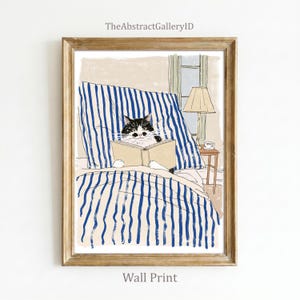 May include: A framed art print featuring a black and white cat reading an open book while lying in a bed with blue and white striped bedding. A small wooden side table with a lamp and a cup sits next to the bed in this illustrated scene.