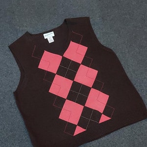 May include: A brown sleeveless sweater with a pink diamond pattern. The sweater has a V-neck and is made of a soft, knitted fabric.