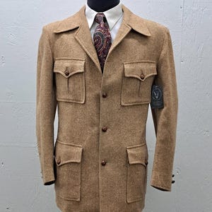 Op de afbeelding: Een bruin tweedjack met een knoopsluiting, vier grote klepzakken en een bruine kraag. Het jack heeft bruine knopen en een bruin-witte paisley-das is zichtbaar onder de kraag.