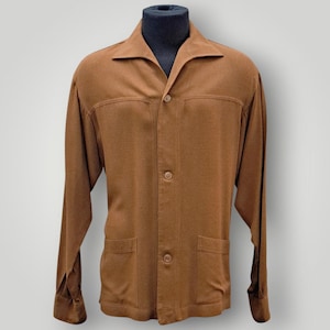 Könnte beinhalten: Ein braunes Langarmhemd mit Kragen und drei Knöpfen. Das Hemd hat zwei Vordertaschen und wird auf einer Schaufensterpuppe präsentiert. Das Hemd ist einfarbig und scheint aus einem weichen Material zu sein.