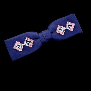 Può includere: Un papillon blu navy con due disegni ricamati a forma di fiocco in rosa e azzurro. Il papillon è annodato al centro e presenta un design semplice ed elegante.