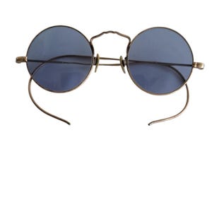 May include: Round eyeglasses featuring circular blue-tinted lenses set within a thin metallic frame. The eyewear includes a curved bridge and long, slender temple arms that hook downward, designed to fit securely around the ears.