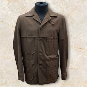 Könnte beinhalten: Braune Jacke mit vier Vordertaschen, Knopfverschlüssen und einem Kragen. Die Jacke wird auf einer schwarzen Schaufensterpuppe präsentiert und hat geknöpfte Manschetten. Das Design der Jacke umfasst eine geknöpfte Klappe auf der rechten Kragenseite.