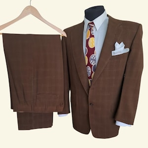 Op de afbeelding: Een bruin geruit pak bestaande uit een blazer en een bijpassende broek. De blazer wordt gedragen over een lichtblauw overhemd met kraag en een donkerrode das met een geel en wit cirkelpatroon. Een wit pochet zit in de zak van de blazer.