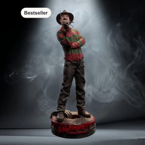 Op de afbeelding: Een beeldje van Freddy Krueger, een horrorfilmkarakter, staat op een voetstuk met de tekst "A Nightmare on Elm Street". Hij draagt een bruine hoed, een rood-groene gestreepte trui en een bruine broek. Het beeldje heeft een "Bestseller" label.
