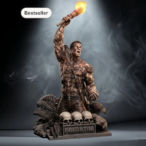 Op de afbeelding: Gedetailleerd beeld van een gespierde figuur die een brandende fakkel omhoog houdt. De figuur is bedekt met camouflage-achtige patronen en staat boven drie schedels. De basis van het beeld luidt "PREDATOR". Een "Bestseller" label staat in de linkerbovenhoek.