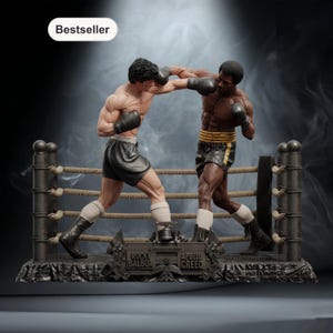 May include: A detailed statue of a boxing match featuring Rocky Balboa and Apollo Creed in a boxing ring. The figures are muscular and wearing boxing gloves and shorts. The ring has ropes and posts, and the base has the names of the boxers.