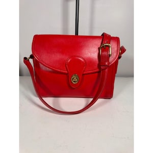 May include: A bright red leather shoulder bag featuring a fold-over flap with a central brass-toned turn-lock closure. The bag includes an adjustable shoulder strap with a brass-toned metal buckle and matching hardware accents on the sides.