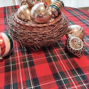 Puede incluir: Una colección de adornos de vidrio vintage en un nido de mimbre marrón. Los adornos son una mezcla de colores y patrones, incluyendo oro, plata, rojo y verde. El nido está sentado sobre un mantel de cuadros rojos y verdes.