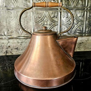 Könnte beinhalten: Ein kupferfarbener Teekessel mit Holzgriff und gebogenem Messinggriff. Der Kessel hat einen Ausguss und einen Deckel. Der Hintergrund ist eine dekorative, gemusterte Wand. Der Kessel wird wahrscheinlich zum Kochen von Wasser für Tee oder andere Heißgetränke verwendet.