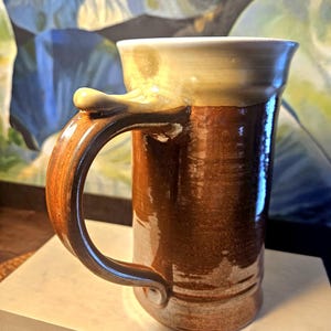 May include: A tall, handcrafted ceramic mug with a large handle. The mug features a brown exterior with a lighter brown and cream-colored rim. The handle is a darker brown, and the mug has a glossy finish. The mug is designed for beverages.