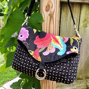 May include: A black crossbody bag featuring a fold-over flap with a vibrant floral pattern in pink, red, yellow, and blue. The main body has a textured grid design with small tan squares. It includes a black strap and two silver metal rings.