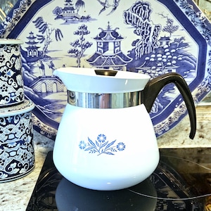 Puede incluir: Cafetera blanca con asa negra y banda plateada. La cafetera presenta un diseño floral azul y se coloca contra una bandeja y botes decorativos con estampado azul y blanco. La cafetera está sobre una vitrocerámica negra.