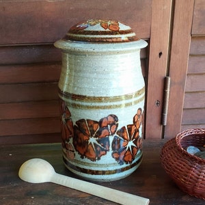 Puede incluir: Un tarro de almacenamiento de cerámica con tapa, con un esmalte marrón y blanco con un patrón de flores naranja y marrón. El tarro está sobre una superficie de madera junto a una cuchara de madera.