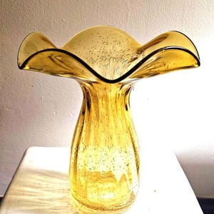 May include: A vintage amber glass vase with a ruffled rim and a flared top. The vase has a bulbous body and a textured surface with small air bubbles. The rim is outlined with a dark color. The vase is sitting on a white surface.