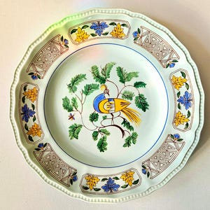 May include: A decorative ceramic plate with a scalloped edge. The plate features a central image of a bird with blue, yellow, and green details, surrounded by a border with floral and geometric patterns. The plate's diameter is approximately 10 inches.